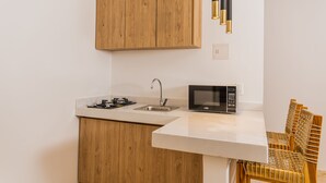 Een kleine keuken met houten kastjes, een magnetron, een spoelbak en een eethoek met een rieten stoel.