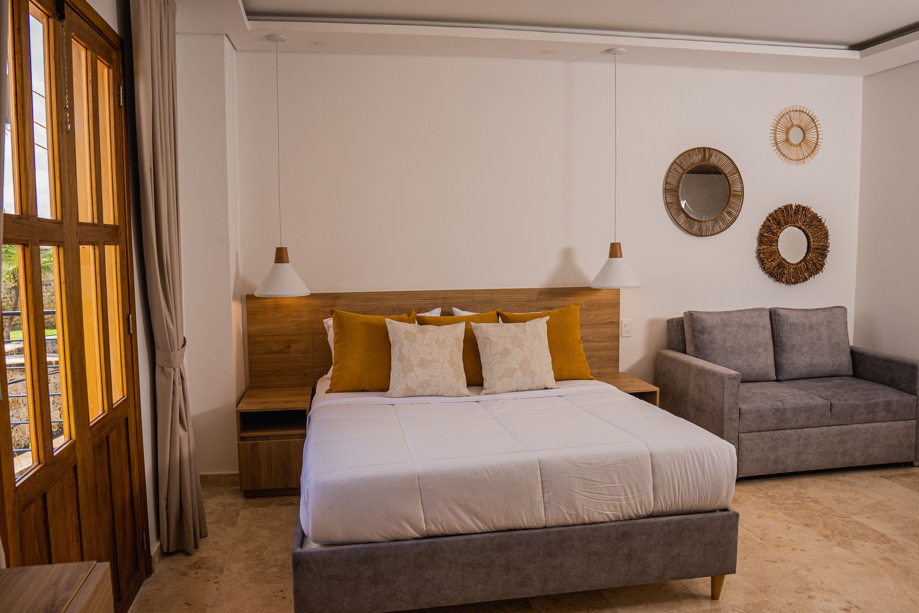 A bedroom with a bed, a grey sofa, a wooden headboard, and two wall mirrors.