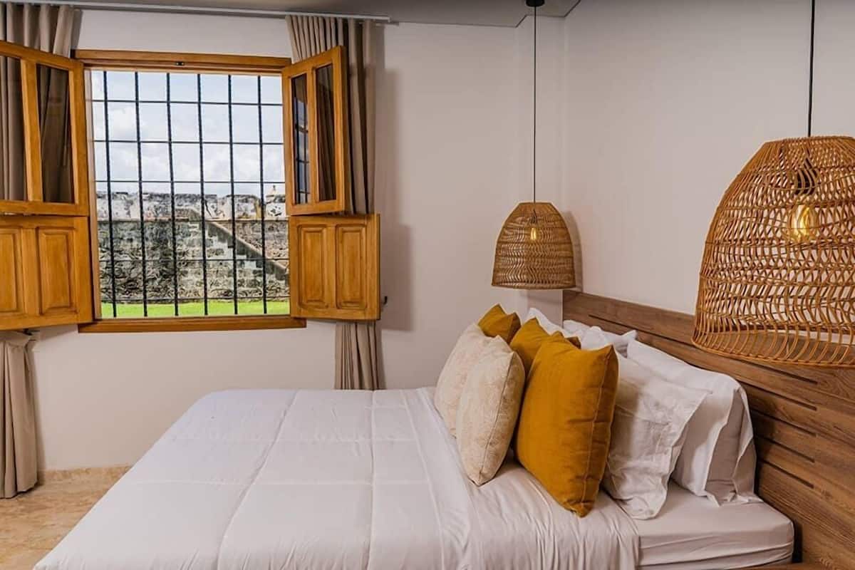 Un dormitorio con una cama, un ventanal con contraventanas de madera, una lámpara de fibras naturales y vistas a un paisaje urbano.
