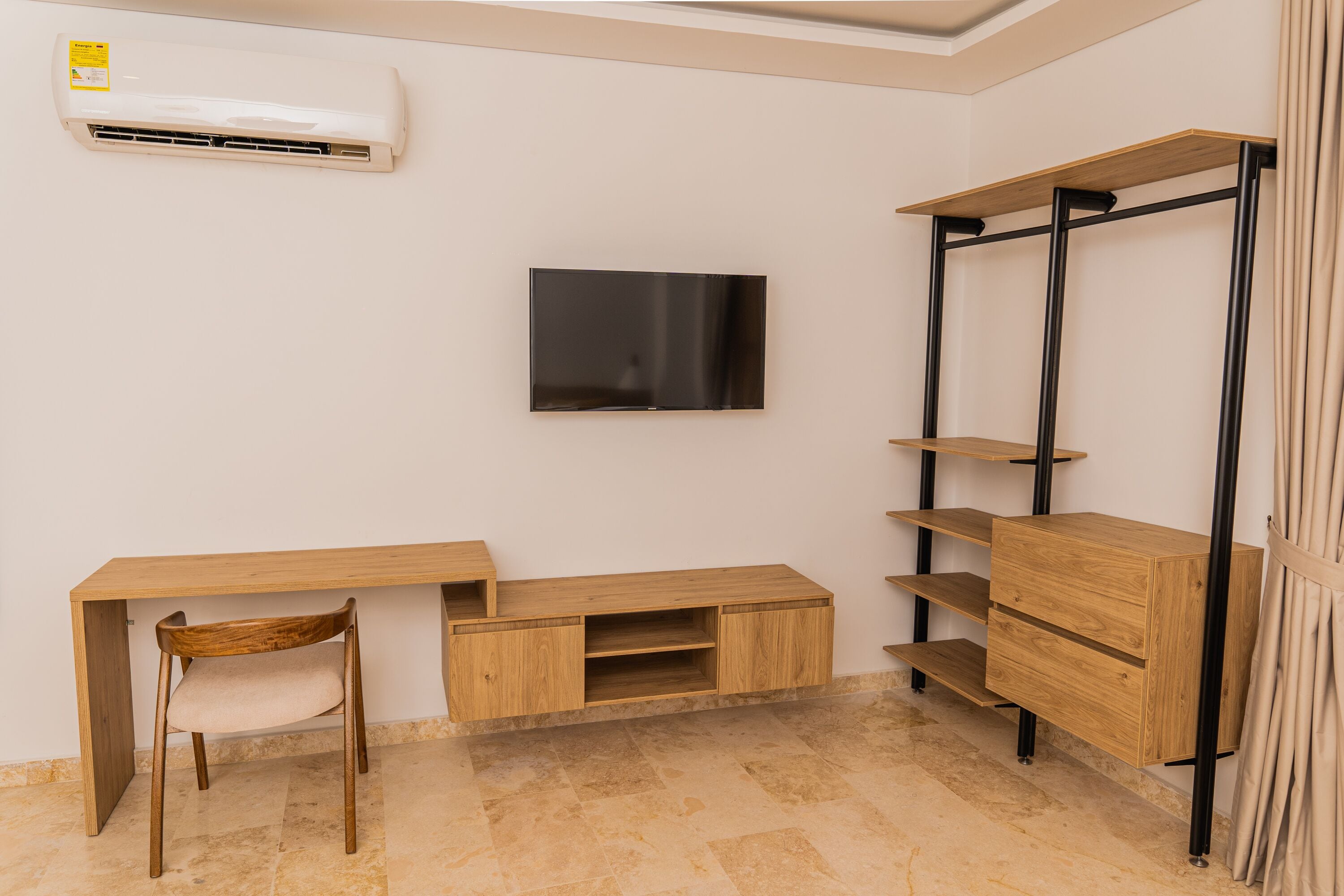 A room with a wooden desk, a chair, a TV mounted on the wall, and a shelving unit.