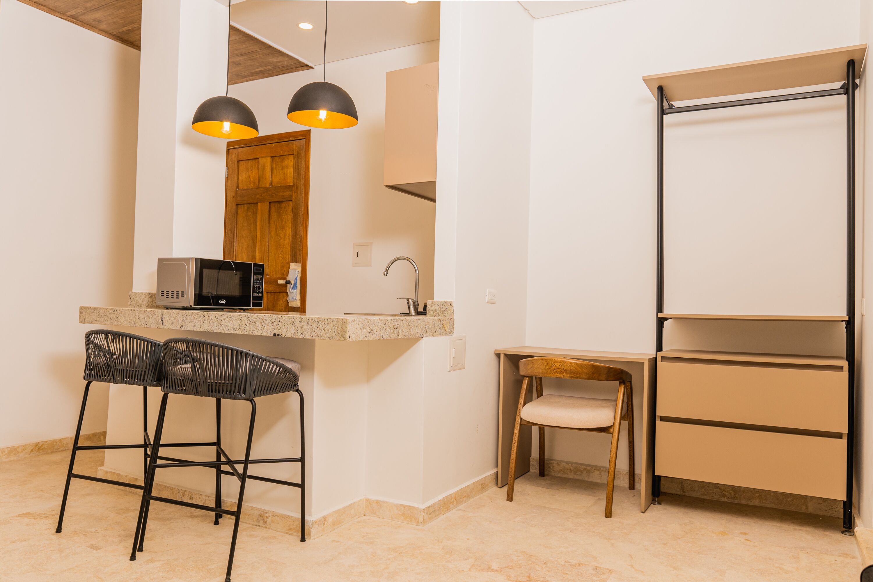 A compact kitchen with a microwave, two bar stools, a wooden chair, and a built-in shelf unit.