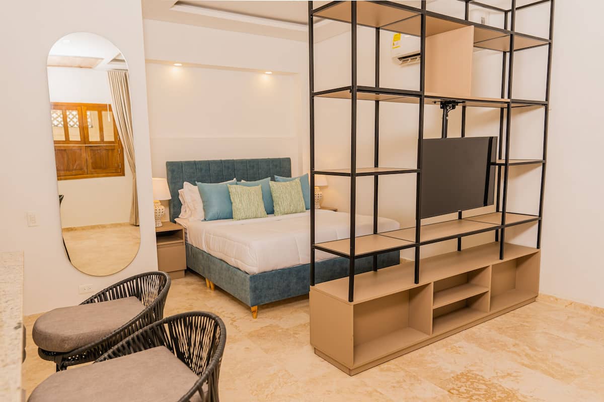 Un dormitorio con cama, televisor en un estante, espejo y dos sillones de mimbre.