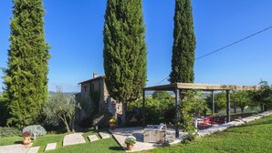 Edifici de pedra amb un pati jardí, alts cipressos i una pèrgola amb seients a l'aire lliure.