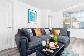 A modern living room with a grey sofa, a coffee table with a bottle, glasses, and fruit, and a framed picture on the wall.