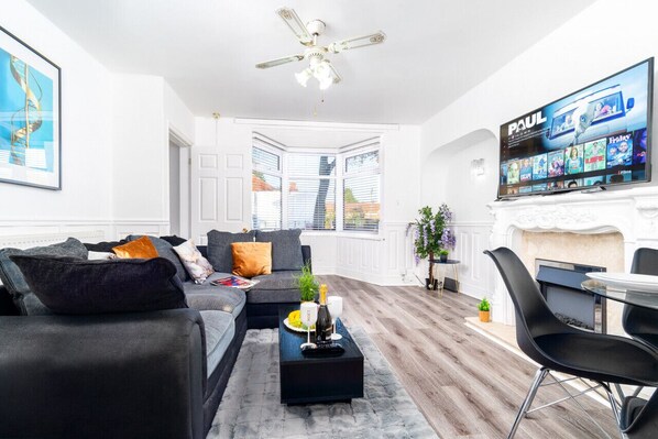 A modern living room with a black sofa, a coffee table with a bottle and glasses, a TV on the wall, and a fireplace.