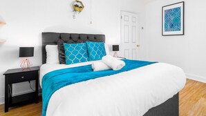 A neatly made bed with a grey headboard, white bedding, and blue and white patterned pillows. There are two bedside tables with lamps, a door, and a framed abstract painting on the wall.