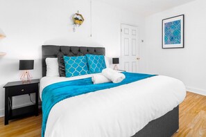 A neatly made bed with a grey headboard, white bedding, and blue and white patterned pillows. There are two bedside tables with lamps, a door, and a framed abstract painting on the wall.