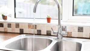 A kitchen sink with a running faucet, a window with a view of greenery, and a tiled backsplash.