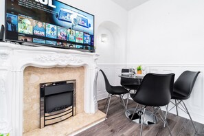A modern living room with a fireplace, a flat-screen TV displaying a movie, and a dining area with black chairs and a small table. - Luxury 3 Bed House -  Garden - Parking (Birmingham)