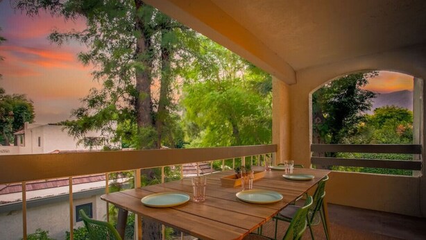 A wooden table with plates and glasses set for two, surrounded by green chairs, on a balcony with a view of trees and a sunset. - Top-floor Condo w/Balcony & Mountain Views (Palm Springs)