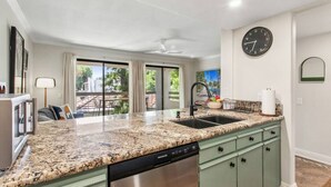 A kitchen with granite countertops, a dishwasher, and a sink. There is a clock on the wall and a window with curtains. - Top-floor Condo w/Balcony & Mountain Views (Palm Springs)