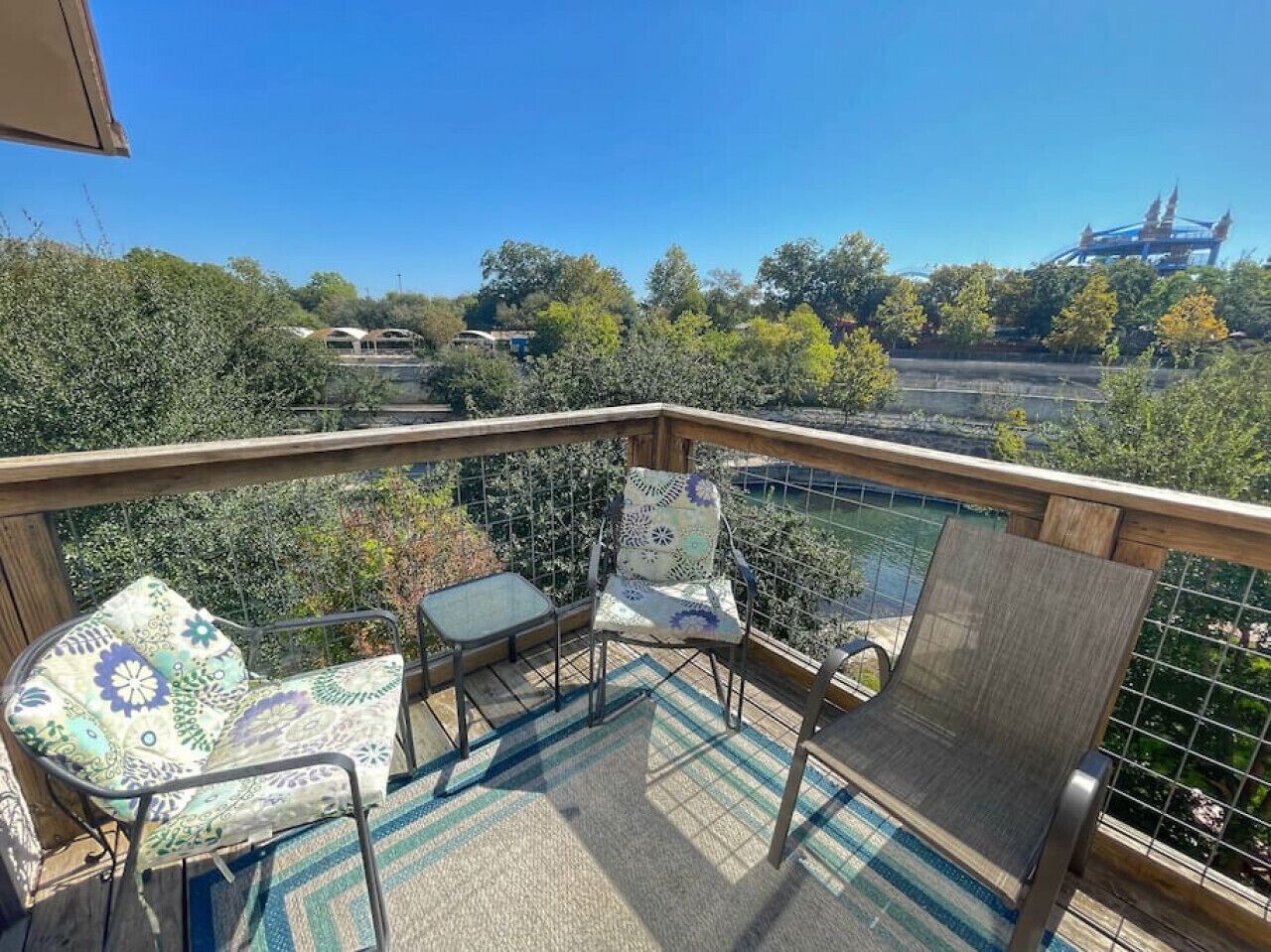 A balcony with three chairs and a table, overlooking a scenic view of trees and a distant bridge.
