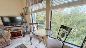 A living room with a TV, a glass-top table, two chairs, a sofa, and a large window with a view of trees.