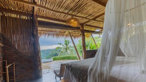 Una habitación con una cama, una escalera de madera, un sillón de mimbre y vistas a la vegetación y una carretera.