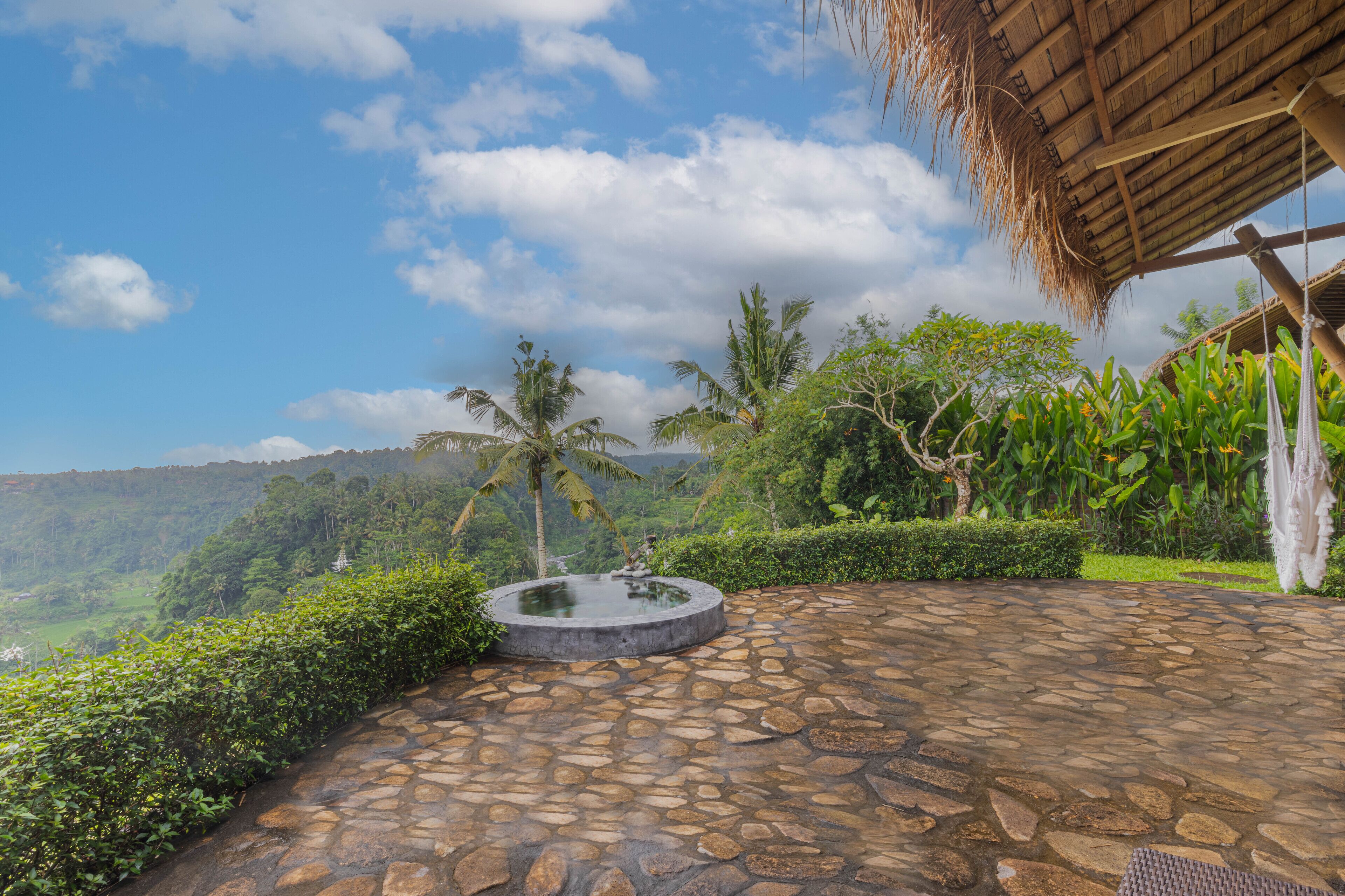 A stone-paved area with a circular water feature, surrounded by lush greenery and a thatched-roof structure.