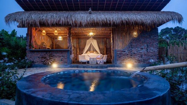 Estructura con techo de paja, jacuzzi en primer plano, cama con dosel y una pequeña mesa con sillas.