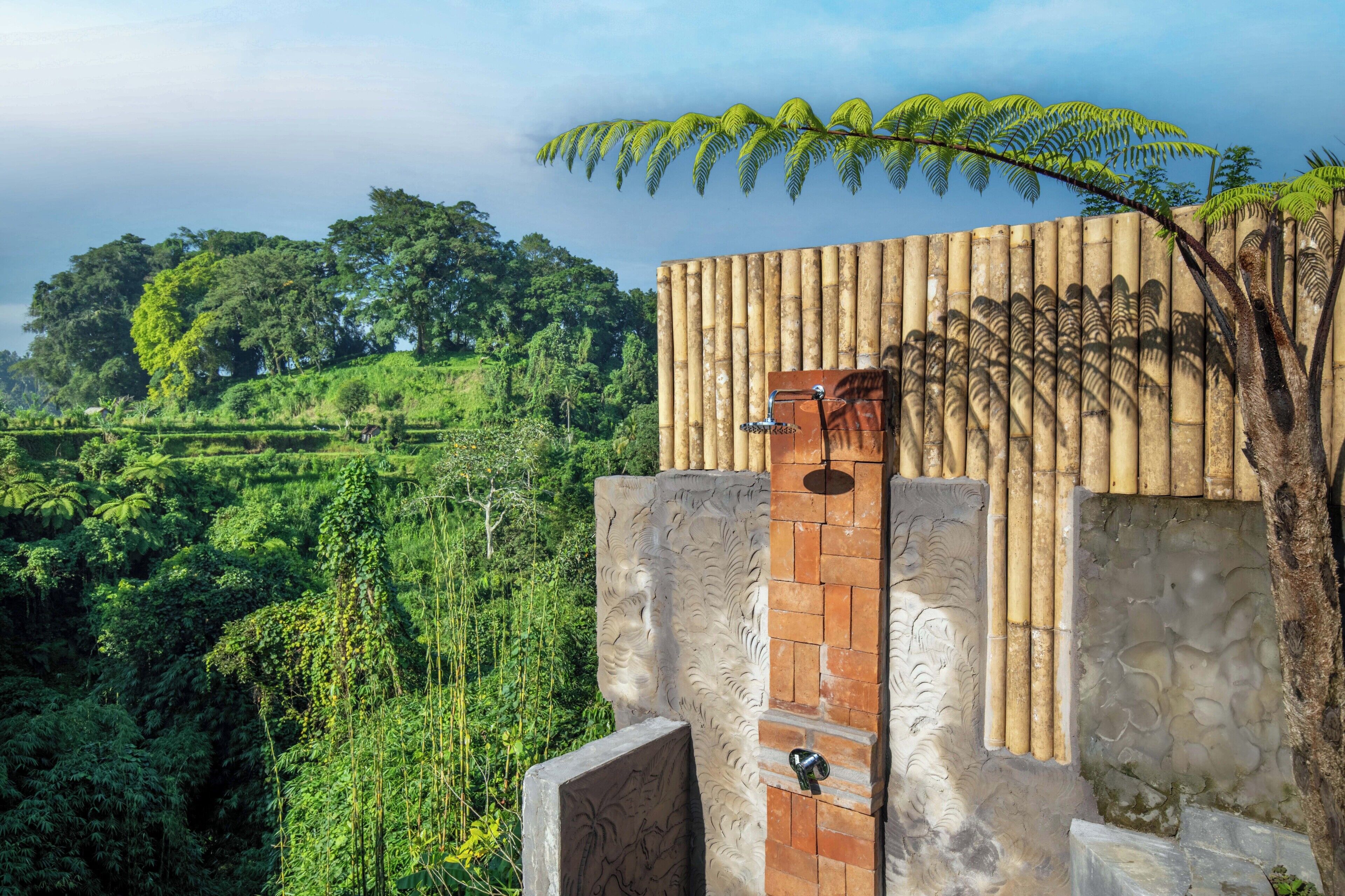 A bamboo wall with a shower area, surrounded by lush greenery and a hill in the background.