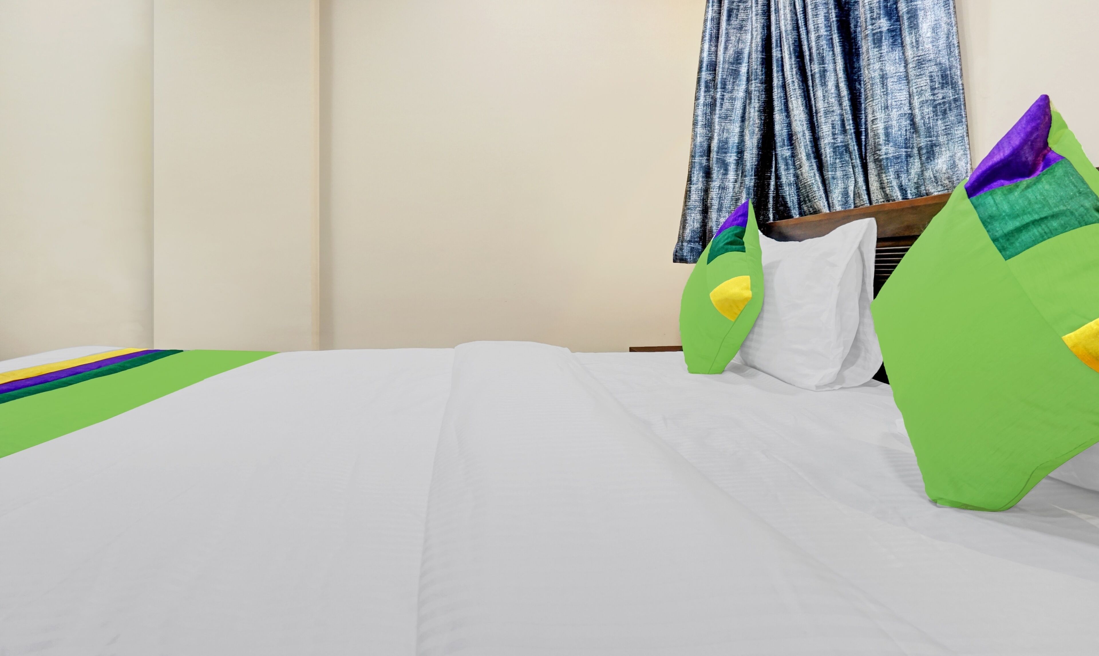 A hotel room with a bed, a blue curtain, and green and yellow pillows.