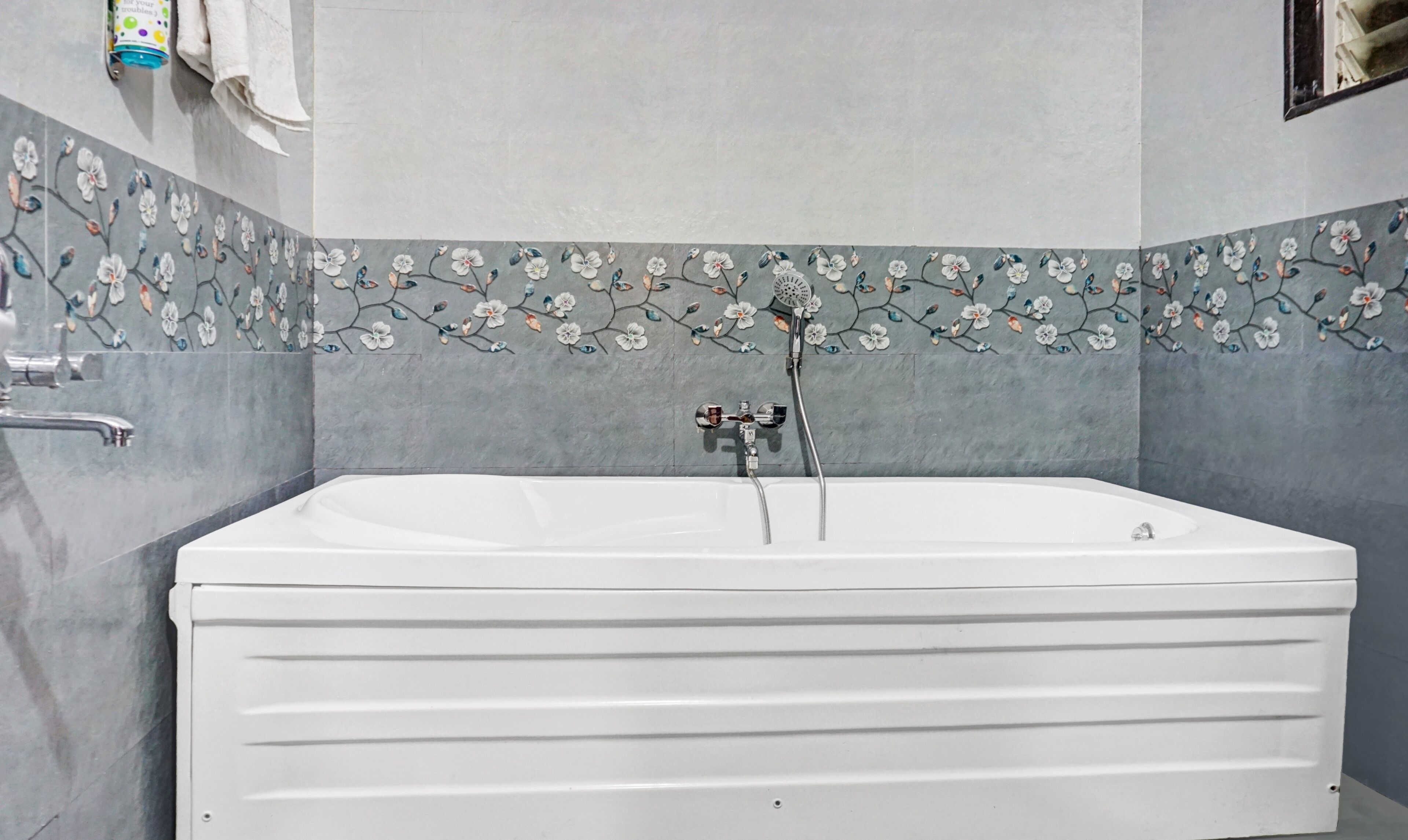 A modern bathroom with a large white bathtub, a shower with a floral tile border, and a towel rack with a bottle.