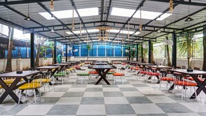 Een ruime, overdekte eetruimte met meerdere tafels en stoelen, een hoge plafond met hangende lampen en grote ramen voor natuurlijk licht.