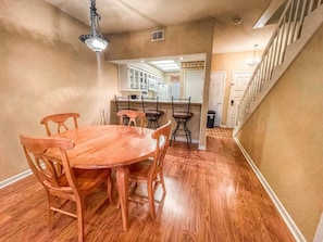 A dining area with a wooden table and chairs, a kitchen in the background, and a staircase leading to another floor.