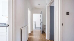 A narrow hallway with white walls and wooden flooring, leading to a door with a blue frame. - The Douglas  2 Bed Apartment in Uddingston (Uddingston)