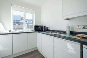 Uma cozinha moderna com armários brancos, fogão a gás, micro-ondas e janela com vista para um prédio.