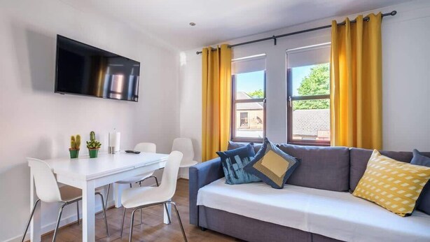A modern living room with a sofa, a dining table with chairs, a flat-screen TV mounted on the wall, and yellow curtains. - The Douglas  2 Bed Apartment in Uddingston (Uddingston)