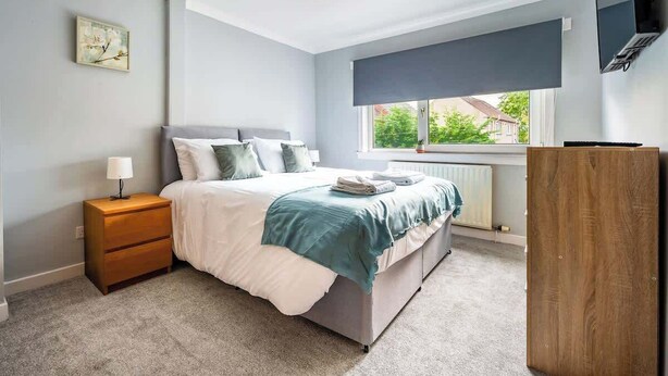 Un dormitorio con cama, mesita de noche, cómoda, ventana y una obra de arte en la pared.