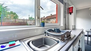 Una cocina con un lavamanos de acero inoxidable, un ventanal con vistas a casas y un letrero rojo de seguridad contra incendios.