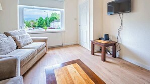 A room with a sofa, a small table, a TV mounted on the wall, and a window with a view of greenery and buildings. - Millfield House -2 Bed in Motherwell near Motorway (North Lanarkshire)