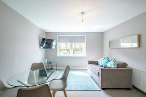 Un salon moderne avec une table Ă manger en verre, un canapĂ© agrĂ©mentĂ© de coussins Ă motifs, un tĂ©lĂ©viseur Ă Ă©cran plat et une grande fenĂȘtre.