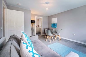 A living room with a grey sofa, a dining table with chairs, a floor lamp, and a painting on the wall.