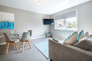 A modern living room with a sofa, a glass dining table, and chairs, a flat-screen TV mounted on the wall, and a window with a view of houses. - Monicas Apartment - 2-Bed Flat in Coatbridge (North Lanarkshire)