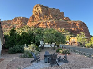 A patio area with Adirondack chairs and a table, surrounded by desert vegetation and a large rock formation in the background. - Red Rock/Mesa Views, No Stairs, Golf, Hike, Relax. EV Charger, Stocked Kitchen! (Sedona)