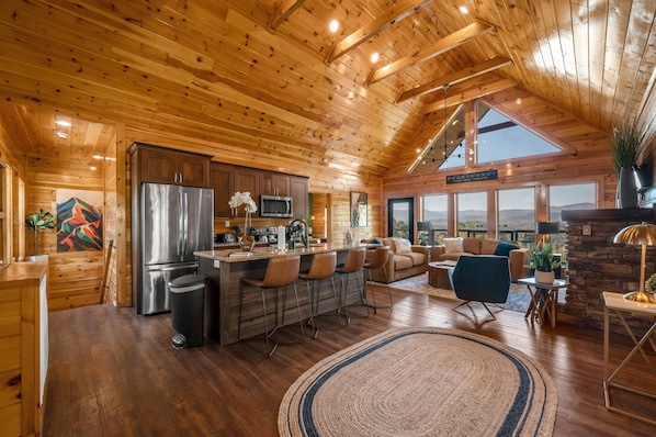 A spacious log cabin with a kitchen featuring a stainless steel refrigerator, a bar area with stools, a stone fireplace, and large windows offering mountain views.