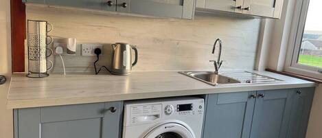 A compact kitchen with grey cabinets, a built-in washing machine, a stainless steel sink, and a kettle.