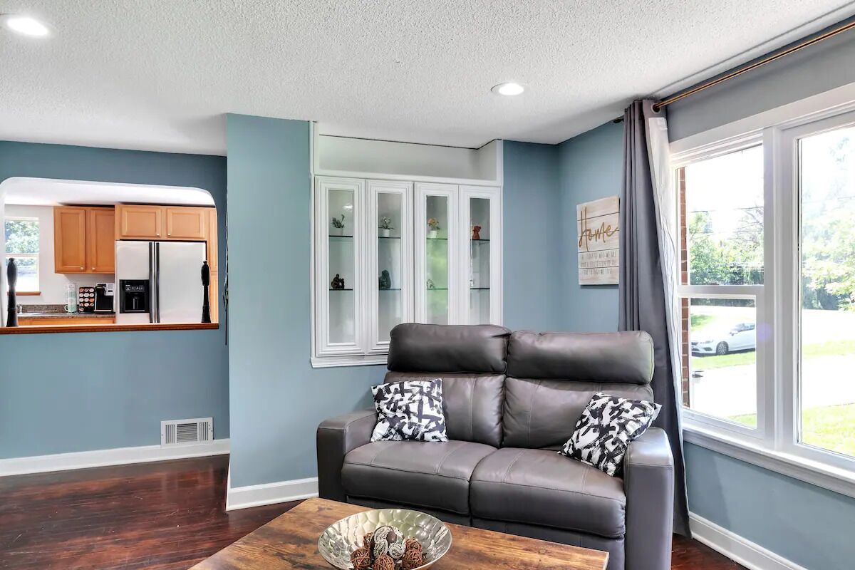 A living room with a grey leather sofa, a wooden coffee table, and a kitchen area with wooden cabinets and a white refrigerator.