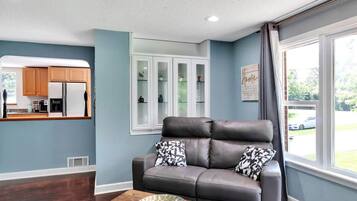 A living room with a grey leather sofa, a wooden coffee table, and a kitchen area with wooden cabinets and a white refrigerator.