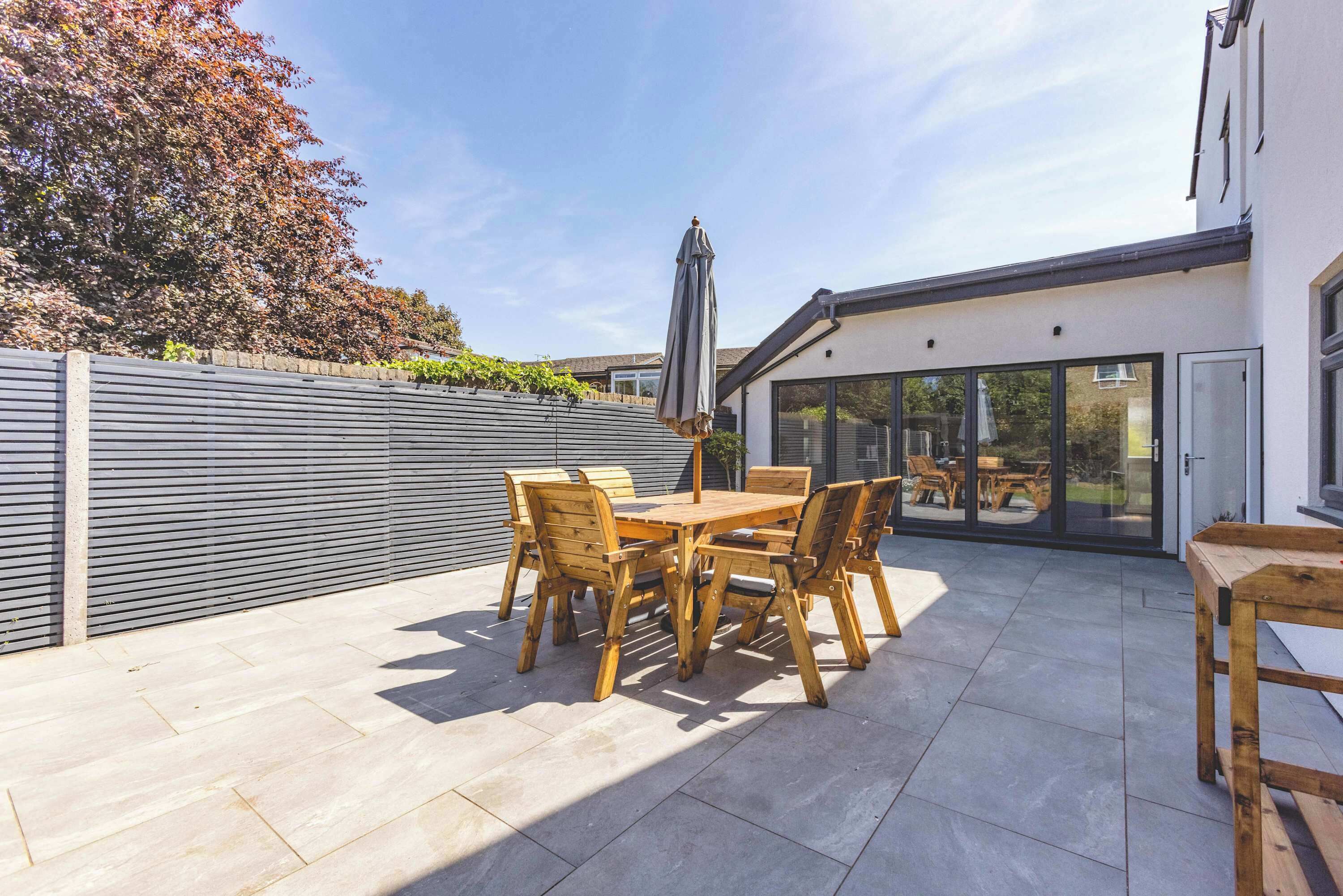 A patio with a wooden table and chairs, a grey umbrella, and a glass door leading to an outdoor dining area.