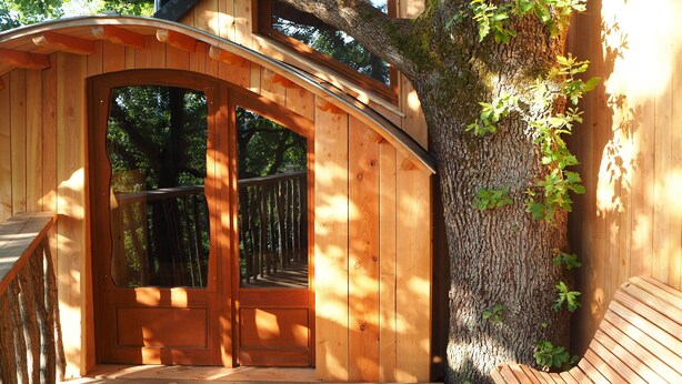A wooden cabin with a curved entrance, large windows, and a staircase leading to an upper level. - Cabane perchée / sauna & cold bath / shared natural swimming pool (L'Isle-Bouzon)
