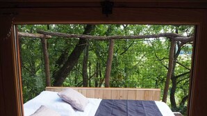 A bed with a view of a forest through a window. - Cabane perchée / sauna & cold bath / shared natural swimming pool (L'Isle-Bouzon)