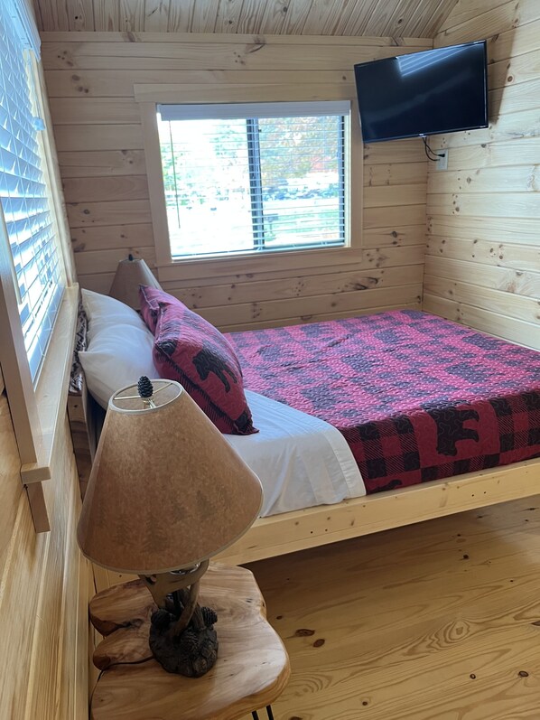 A wooden cabin room with a bed, a mounted TV, and a window with blinds. - Cozy Log Cabin w/ Loft #14. Minutes from Seven Springs and Hidden Valley! (Champion)