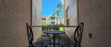 An outdoor balcony with a round table and two chairs, overlooking a building with a tiled roof and palm trees.