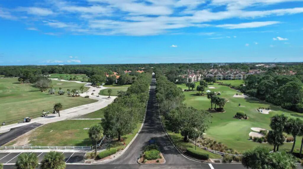 En vy över en golfbana tagen från luften, med en väg som går genom mitten, omgivande grönska och bostadshus i bakgrunden.