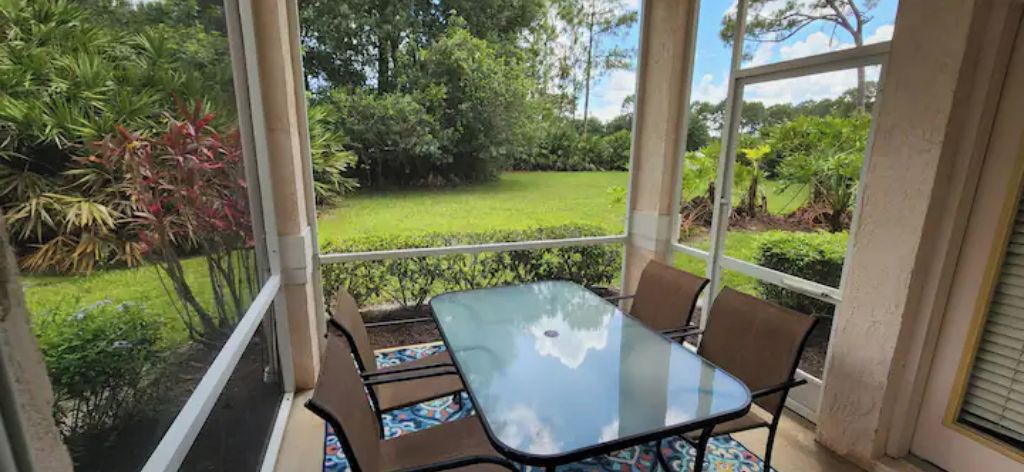 A glass-enclosed patio with a table and chairs, overlooking a well-maintained garden with various plants and a lawn.
