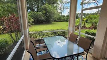 A glass-enclosed patio with a table and chairs, overlooking a well-maintained garden with various plants and a lawn.