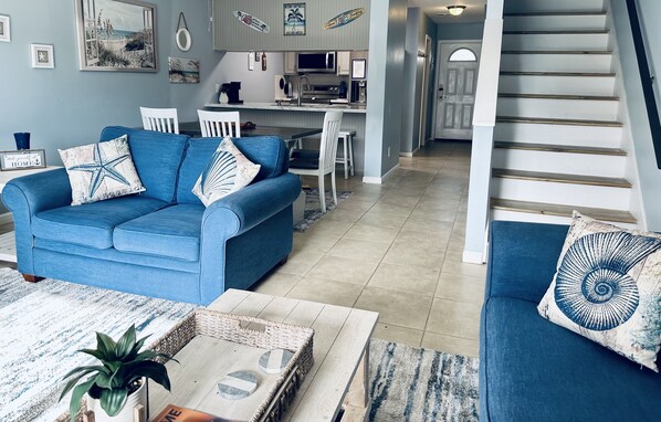 A living room with a blue sofa, white chairs, a coffee table, and a kitchen in the background.
