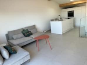 Un salon moderne avec un canapé gris, un petit tabouret rouge et un coin cuisine doté de meubles blancs et d’un four encastré.