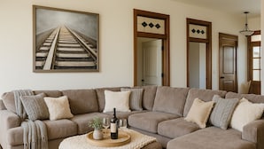 A living room with a sectional sofa, a coffee table with wine glasses and a bottle, and a framed picture of a railway track. - Hot Tub + 1,600 sq ft Golf-Course Patio • Game Room & Luxury Living (Billings)